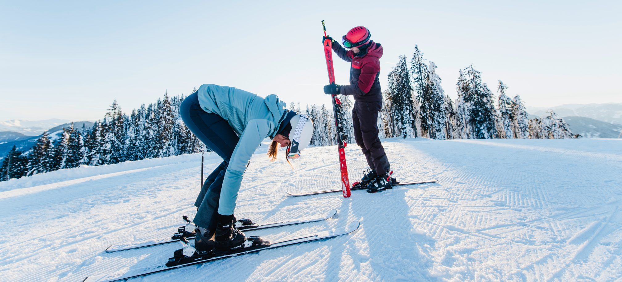Eine Skifahrerin beugt sich zu ihren Schnellen bei den Skischuhen hinunter und der Skifahrer schaut sich gerade den abgeschnallten Ski an während er mit dem anderen noch auf der Piste steht