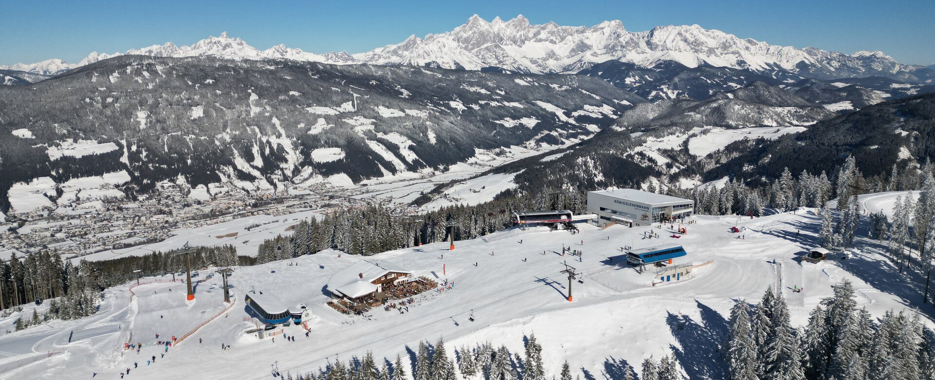 Eine Gondelstation, zwei Sesselliftstationen und eine Skihütte von oben und im Tal unten sind viele Häuser zu sehen