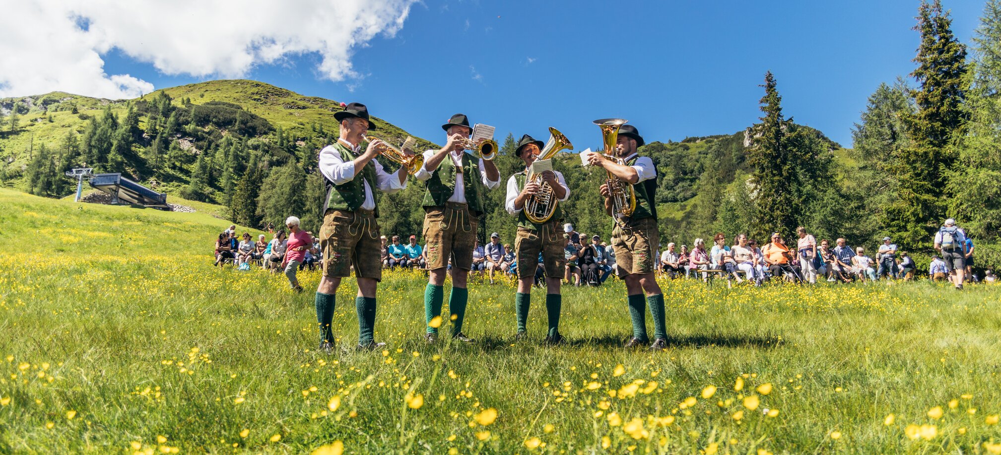 4 musicians are standing on a meadow playing brass instruments and many people are sitting in the background | © Reiteralm