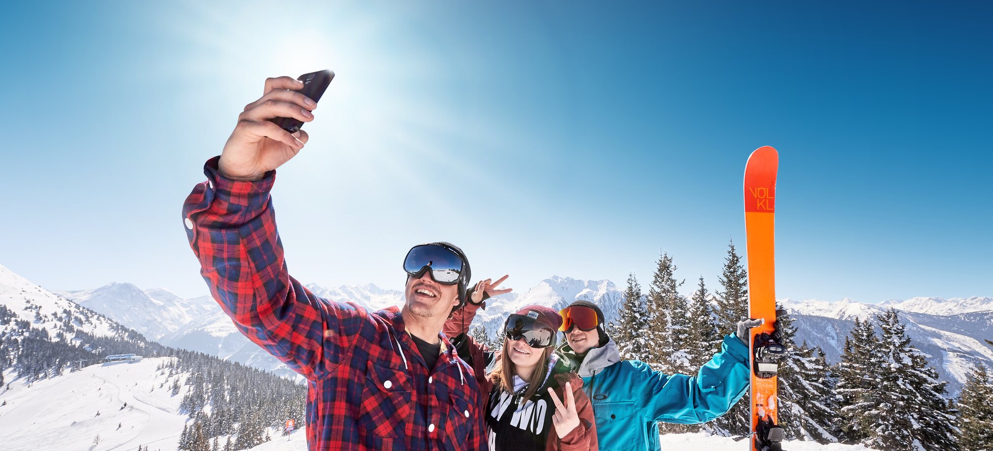 Three teenagers in ski goggles take a selfie on the snow-covered mountain and the one in the back holds an orange ski