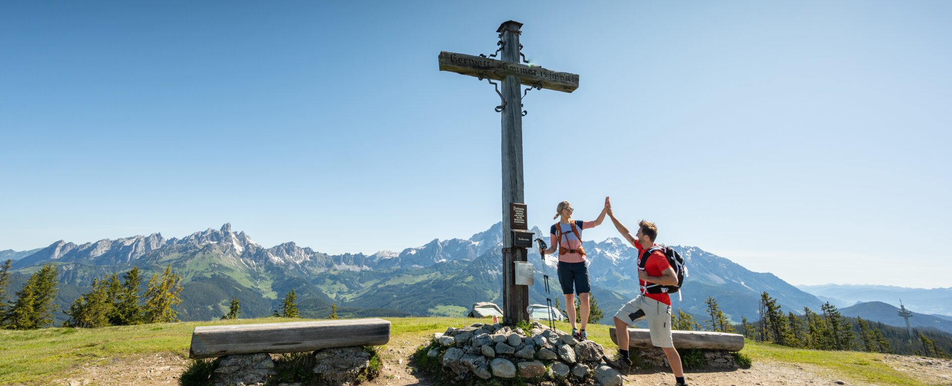 Two people stand on a plateau by a summit cross and give each other a high five | © Tourismusverband Radstadt/Lorenz Masser