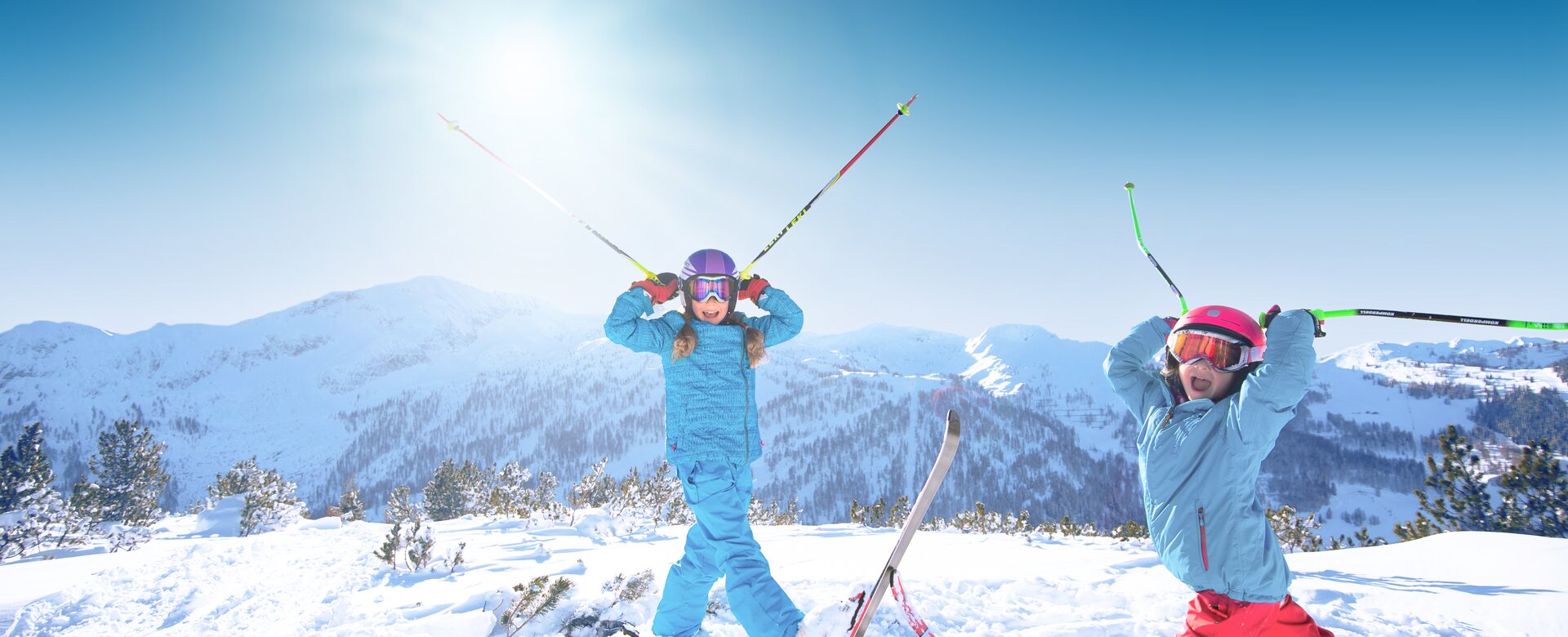 Zwei Kinder stehen im Tiefschnee und halten sich die Skistöcke an den Helm, so dass sie aussehen wie Hörner, und haben den Mund weit offen