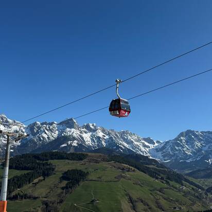 Rote Gondel der Abergbahn schwebt im Sommer vor grüner Landschaft und schneebedecktem Hochkönig-Massiv bei blauem Himmel. | © Aberg - Hinterthal - Bergbahnen AG