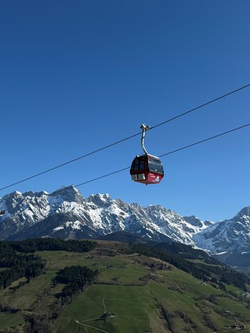 Rote Gondel der Abergbahn schwebt im Sommer vor grüner Landschaft und schneebedecktem Hochkönig-Massiv bei blauem Himmel. | © Aberg - Hinterthal - Bergbahnen AG