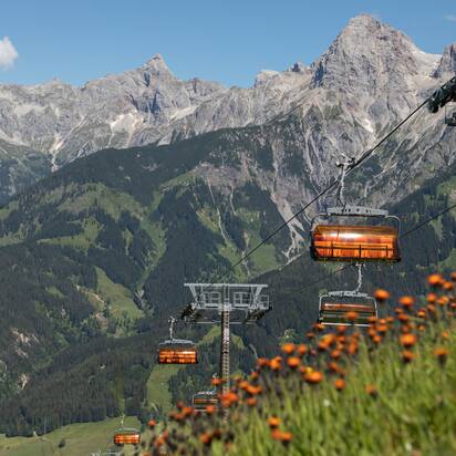 Orange Sessellifte der Hochmaisbahn fahren im Sommer vor alpiner Kulisse des Hochkönigs über blühende Wiesen. | © Roland Haschka
