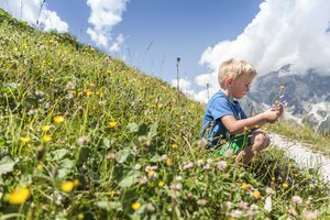 Kleiner Junge sitzt auf einer Almwiese und pflückt bunte Blumen vor imposanter Bergkulisse unter blauem Sommerhimmel. | © Felsch Fotodesign