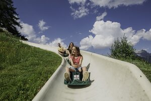 Mädchen rodelt mit zwei Erwachsenen hinter sich die Sommerrodelbahn Biberg hinunter, grüne Wiese, blauer Himmel. | © Hochkönig / Roland Haschka