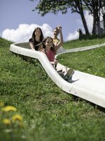 Lachendes Mädchen fährt mit Familie auf Sommerrodelbahn den grünen Hang hinunter, Sommerwiese im Vordergrund. | © Hochkönig / Roland Haschka