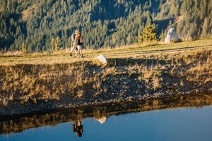 Zwei Wanderer gehen am Ufer eines kleinen Teichs bei der Bürglalmbahn in Dienten über eine Wiese. | © Hochkönig Bergbahnen