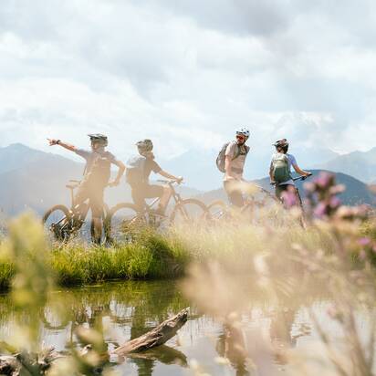 Vier Mountainbiker stehen mit Rädern am Ufer eines kleinen Teichs vor Bergen bei der Bürglalmbahn in Dienten | © Hochkönig Bergbahnen