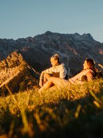 Zwei Wanderinnen sitzen im Abendlicht auf einer Wiese in Dienten vor einem felsigen Bergmassiv nahe der Gabühelbahn. | © Hochkönig Bergbahnen
