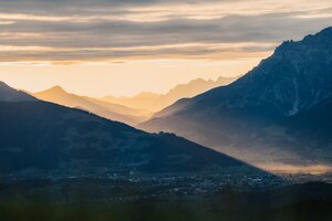 Blick über Dienten auf ein Tal mit Bergsilhouetten, Sonnenstrahlen und dicht bewölktem Himmel im Morgenlicht. | © Hochkönig Bergbahnen