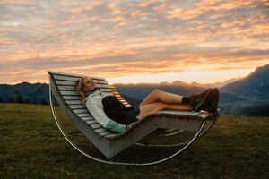 Eine Frau liegt auf einer hölzernen Liegebank auf einer Wiese in Dienten mit Bergpanorama bei Sonnenuntergang. | © Hochkönig Bergbahnen