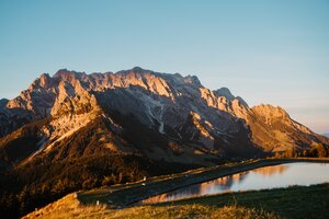 Sonnenlicht beleuchtet ein felsiges Bergmassiv über einem Speicherteich in Dienten unter klarem Himmel. | © Hochkönig Bergbahnen