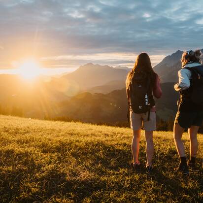 Zwei Wanderinnen stehen auf einer Wiese in Dienten und blicken bei Sonnenuntergang auf Berge nahe der Gabühelbahn. | © Hochkönig Bergbahnen