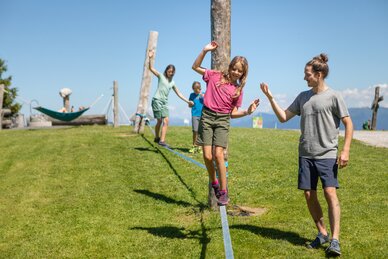 Ein Mädchen balanciert auf einer Slackline in Mühlbach, während ein Mann hilft und zwei Kinder folgen. | © Hochkönig Bergbahnen, Roland Haschka