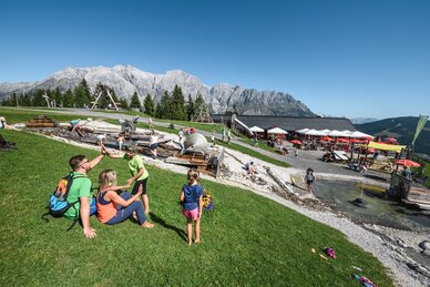 Familien genießen den Wasserspielplatz an der Karbachalm vor imposanter Hochkönig-Kulisse bei Sonnenschein