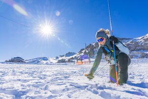 Frau kniet im Schnee mit Sonde und LVS-Gerät, Sonnenschein und Berge im Hintergrund.  | © René Eduard Perhab