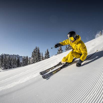 A skier in yellow outfit speeds down a freshly groomed slope, carving turns as snow sprays into the air. | © Mirja Geh