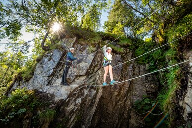 Zwei Kinder mit Helm und Klettergurt auf Seilbrücke an Felswand, sonniges Wetter, Bäume und Felsen im Hintergrund. | © Snow Space Salzburg, Christoph Huber