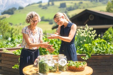 Zwei Frauen begutachten Kräuter an einem Tisch mit Gläsern und Korb vor Hochbeeten, Haus und grünen Hängen. | © Hochkönig Tourismus GmbH