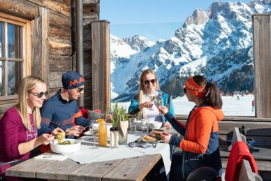 Four people dining and drinking on wooden terrace of mountain hut with Alpine view | © Hochkönig Tourismus GmbH