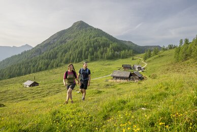 Two hikers walk across an alpine meadow near Altenmarkt, with wooden huts and a forested mountain. | © Lorenz Masser