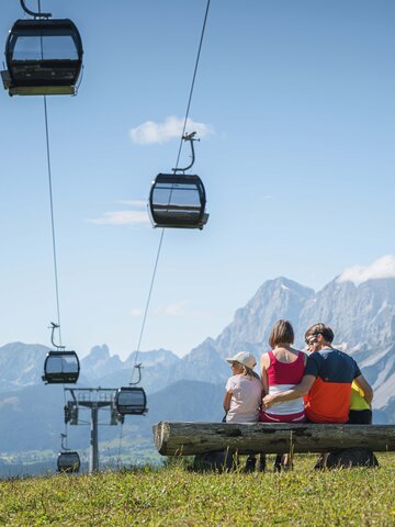 A family sits on a wooden bench under gondolas, admiring the impressive mountain panorama in the Austrian region of Ski amadé.