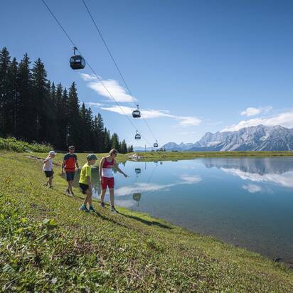 Eine Familie spaziert entlang eines spiegelklaren Bergsees unter schwebenden Gondeln mit Aussicht auf das Dachsteinmassiv.