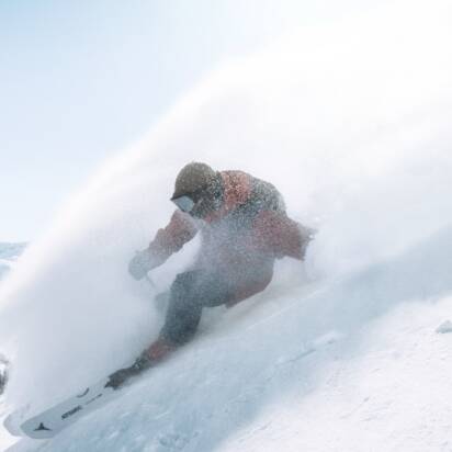 Snowboarder fährt im Tiefschnee eine steile Hangflanke hinab, Pulverschnee spritzt auf