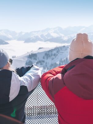 Two people in winter clothes stand on terrace overlooking snowy mountains with skis in the foreground