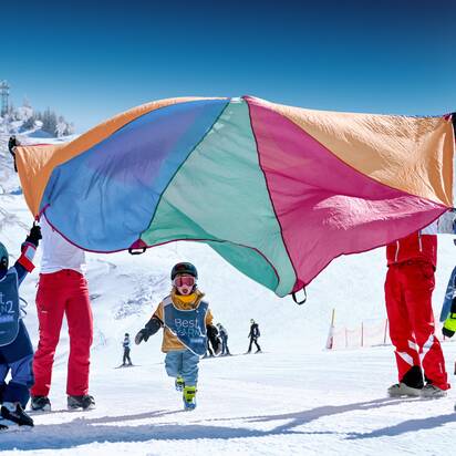 Children in Best Learn2Ski vests play under colourful parachute on snowy slope with ski instructors in the background