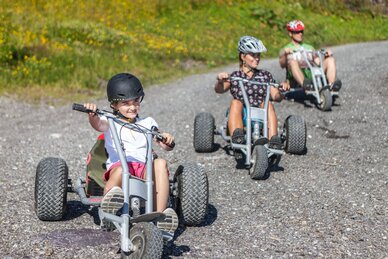 Kind und Erwachsene fahren mit Helmen auf Mountaincarts über eine Schotterstraße in sommerlicher Berglandschaft