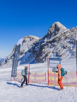 Zwei Wintersportler vor einem Trainingsbereich im Schnee mit Fahnen, Zaun und Gebirgskulisse am Dachstein.  | © René Eduard Perhab