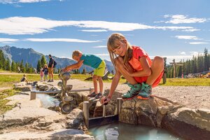 Zwei Kinder spielen an einer Wasserspielanlage mit Rinnen und Schleusen im Hopsiland Planai, Bergkulisse im Hintergrund. | © Christine Höflehner