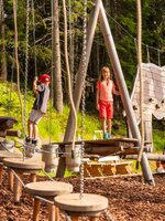 Zwei Kinder balancieren im Klettergarten von Hopsiland Planai auf Holzelementen mit Seilen und Figuren im Wald. | © Christine Höflehner