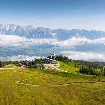 Weite Alm mit Bergstation und Aussichtsturm, umgeben von Wiesen, Wald und Bergketten, teils in Wolken gehüllt. | © Josh Absenger