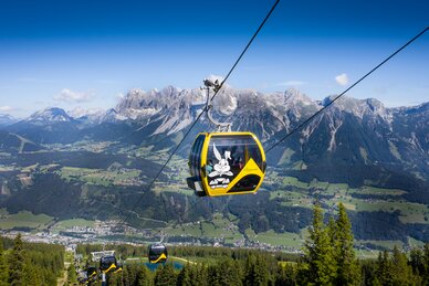 Gelbe Gondel mit Hopsiland-Motiv über der Planai mit Blick auf das Dachsteinmassiv und das grüne Ennstal im Sommer. | © Josh Absenger