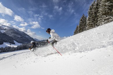 Two skiers descend through deep powder snow in sunshine near forest edge | © filzmoos.ski