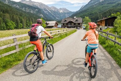 Erwachsener und Kind fahren auf Mountainbikes durch ein grünes Tal mit Holzzäunen und Bergen im Hintergrund. | © Shuttleberg