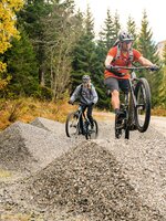 Zwei Mountainbiker fahren über Kieshügel im Wald, einer springt mit dem Bike. | © Shuttleberg