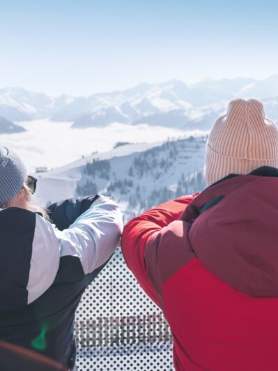 Two people in winter clothes stand on terrace overlooking snowy mountains with skis in the foreground
