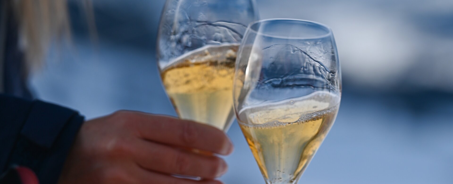 Zwei Sektgläser beim Anstoßen, im Hintergrund verschneite Berge und blauer Himmel