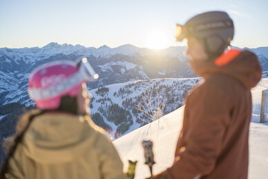 Two people in ski gear with helmets and goggles watching sunset over snowy mountains in Dorfgastein | © Dorfgasteiner Bergbahnen