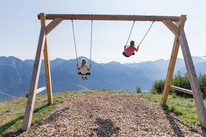 Zwei Kinder schaukeln auf Holzschaukel vor Bergpanorama mit Fernblick über das sonnige Gasteinertal | © Gasteiner Bergbahnen AG