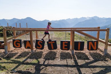 Mädchen balanciert über Spielgerät mit GASTEIN-Buchstaben vor Bergpanorama im Gasteinertal | © Gasteiner Bergbahnen AG