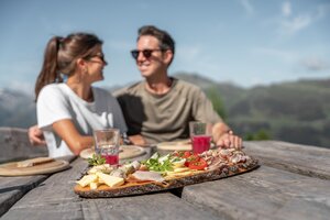 Paar genießt Brettljause mit Käse, Speck und Saft auf Holztisch vor Bergpanorama | © Skigastein, seven frames