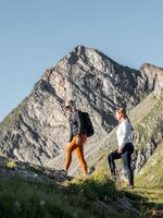 Zwei Frauen wandern am Graukogel bergauf, im Hintergrund ein markanter, felsiger Berggipfel bei klarem Himmel | © Skigastein, seven frames