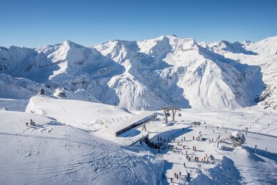  The top station of a cable car from above, with skiers next to it, surrounded by snow-covered mountains.