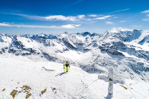  A freeskier stands next to a frosty summit cross, holding the skis in one hand and the poles in the other. | © Christoph Oberschneider